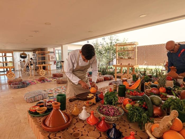 Un cocinero preparando tajines rodeado de cestas de productos y especias marroquíes.
