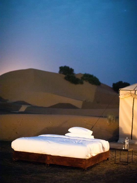 Una cama preparada bajo el cielo abierto junto a una tienda del desierto al anochecer.